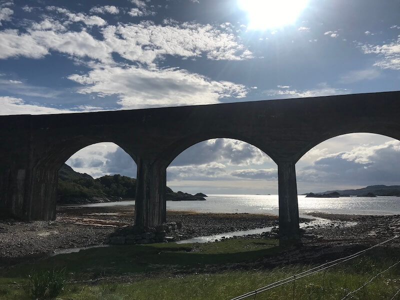 A tall stone railway viaduct with multiple arches stands silhouetted against the sunlit sky, framing views of a rocky shoreline and the glistening sea beyond. The scene is peaceful with scattered clouds, green grass in the foreground, and soft sunlight reflecting off the water.