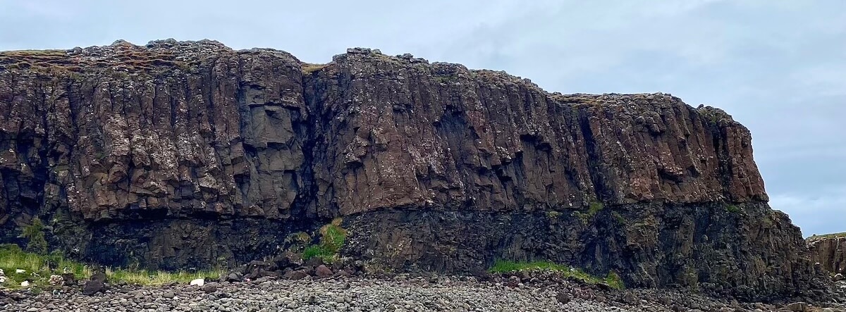 A striking basalt rock formation on Sanday, Isle of Canna, showcasing rugged, vertical columns that rise dramatically from a stony shoreline. The dark, weathered texture of the rock contrasts against a cloudy sky, highlighting the raw natural beauty of this coastal landscape.