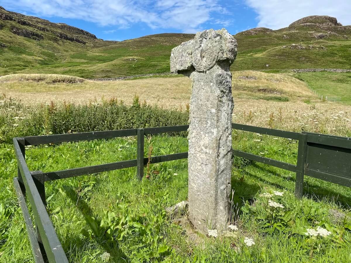 An ancient Celtic cross on the Isle of Canna, standing within a simple wooden fence amidst lush green grass. The stone cross, weathered by time, is set against a backdrop of rolling hills and a bright blue sky with wispy clouds
