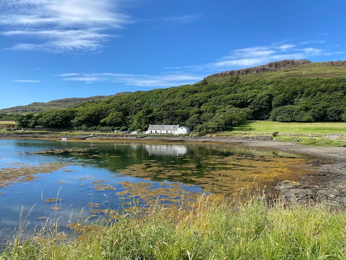 A picturesque view from the harbour on the Isle of Canna, featuring a quaint white cottage nestled by the water's edge. The scene includes a calm bay reflecting the clear blue sky and surrounding greenery, with rolling hills and basalt cliffs in the background.
