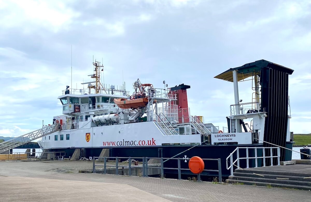 The 'Loch Nevis' ferry docked at the Isle of Canna harbour, operated by Caledonian MacBrayne. The vessel features a white hull with red and black accents, and visible lifeboats on deck, under a partly cloudy sky. The CalMac website address is displayed prominently on the side of the ferry