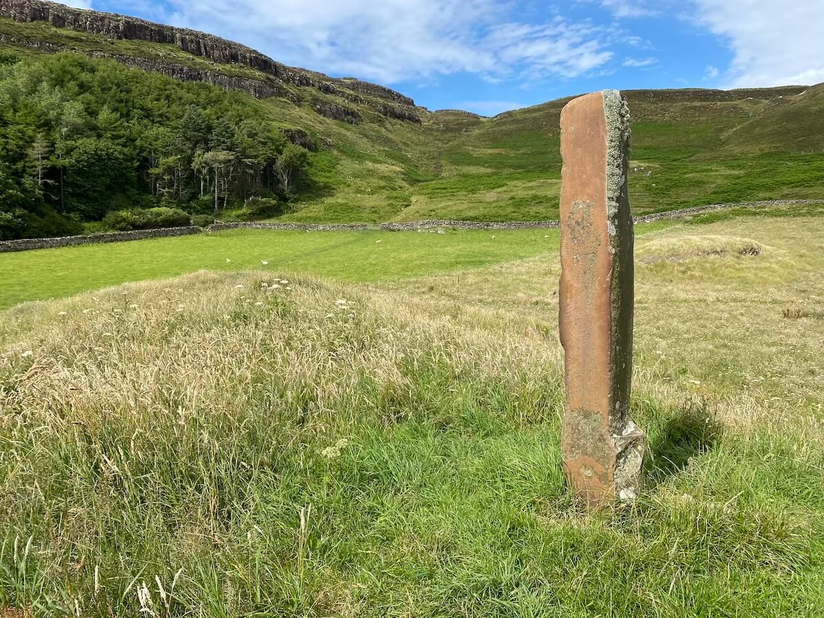 "The Punishment Stone on the Isle of Canna, a tall, narrow standing stone set in a grassy field. The weathered stone stands against a backdrop of rolling green hills, dense woodland, and a bright blue sky with scattered clouds