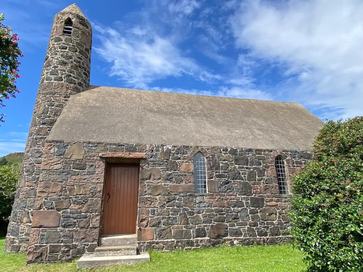 Rhu Church on the Isle of Canna, a quaint stone building featuring a tall, narrow tower and arched windows. The church is constructed from multi-coloured stone and has a simple, rustic appearance, set against a bright blue sky with scattered clouds and surrounded by greenery
