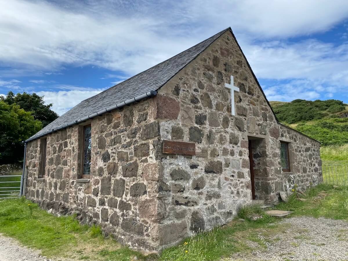 St Columba's Church on the Isle of Canna, a small rustic chapel constructed from rough-hewn stones. The building features a simple white cross on the front gable, stained glass windows, and a slate roof, set against a backdrop of green hills and a blue sky with scattered clouds