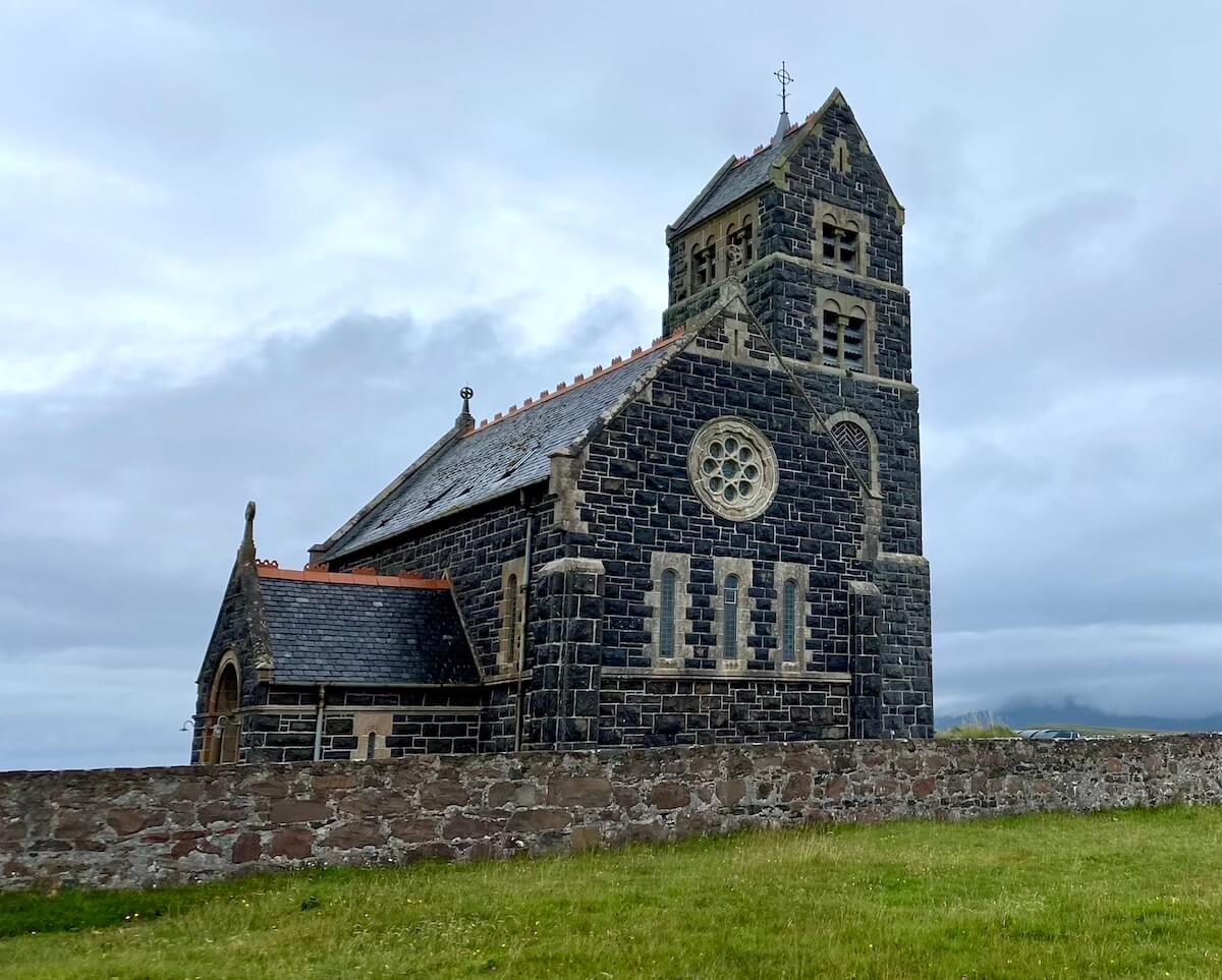 St Edward's Church on Sanday, Isle of Canna, a striking structure built from dark stone with lighter stone accents. The church features a tall tower with arched windows and a rose window on the main facade. The building is surrounded by a low stone wall and sits against a backdrop of a cloudy sky