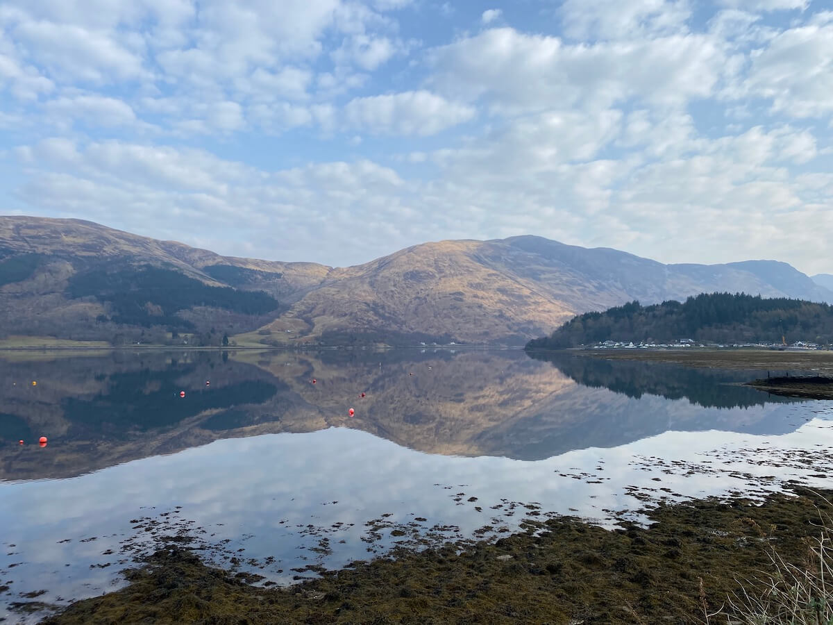 loch in early spring with stark trees and reflection of mountains on still water