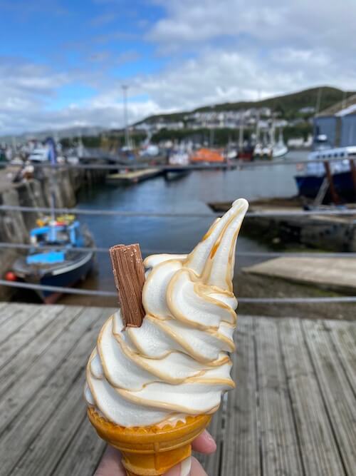 a whipped ice cream in a cone with Mallaig harbour in the background