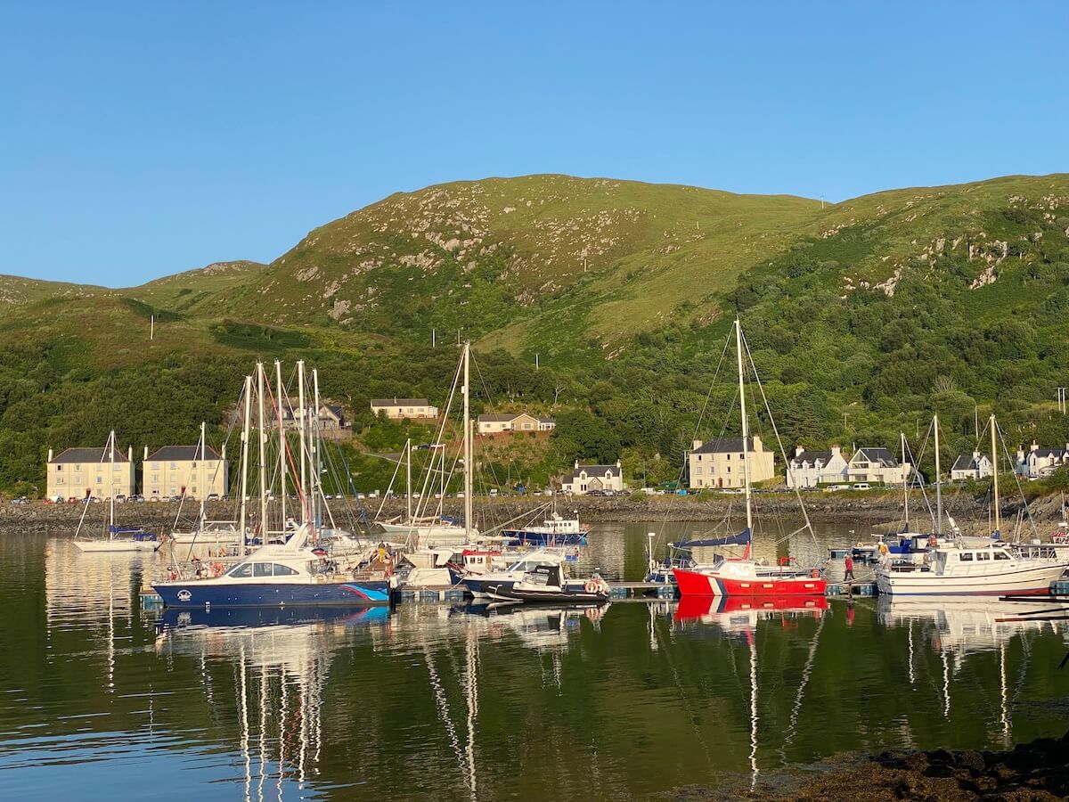 still waters in mallaig harbour with sailing boats, green hills and houses in the background
