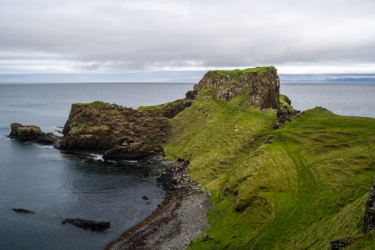 The dramatic coastline of Brother's Point on the Isle of Skye, showcasing lush green grassy terrain and rocky cliffs against the backdrop of a calm sea. The patterns of natural erosion are visible on the slopes, and a pebbly beach curves along the water's edge under a cloudy sky