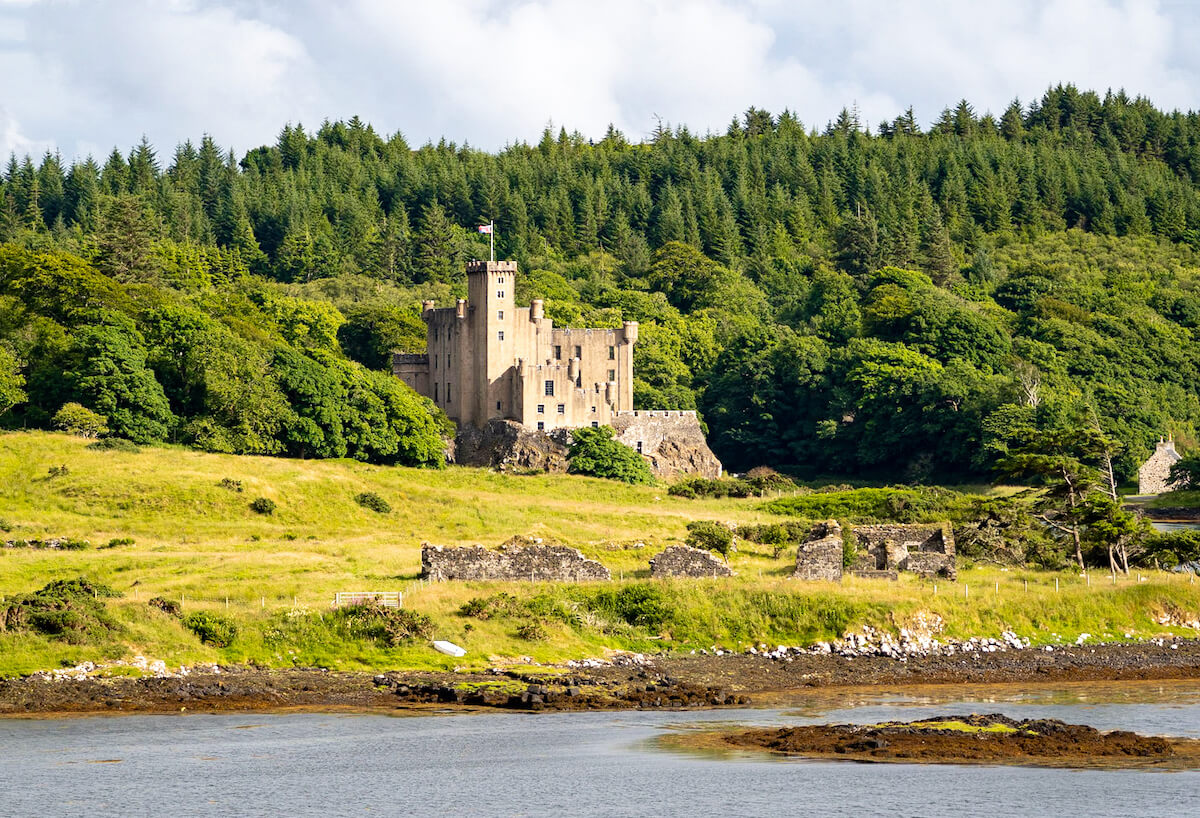 Dunvegan Castle, a stately home with a robust and storied silhouette, stands proudly amidst lush greenery on the Isle of Skye. Its historic stone façade contrasts with the dense forest backdrop, while the calm waters of Loch Dunvegan reflect a peaceful Scottish landscape