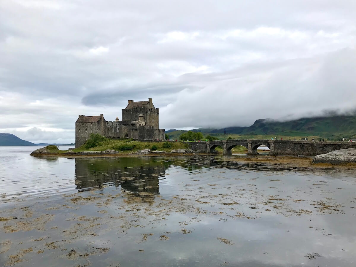Eilean Donan Castle, a majestic 13th-century fortress, sits on a small tidal island where three sea lochs meet, under a heavy grey sky. Its reflection is mirrored in the calm waters, connected to the mainland by a graceful stone bridge, set against the backdrop of Scotland’s misty mountains