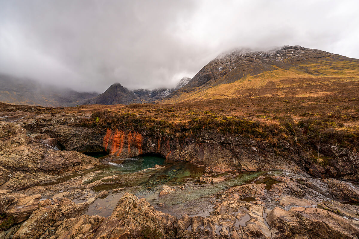 A rugged landscape at the Fairy Pools on the Isle of Skye, with a foreground of twisted rock formations and a serene pool of water, reflecting hints of orange from the iron-rich soil. Misty mountains rise in the background, their peaks shrouded in clouds, conveying the raw beauty of the Scottish Highlands