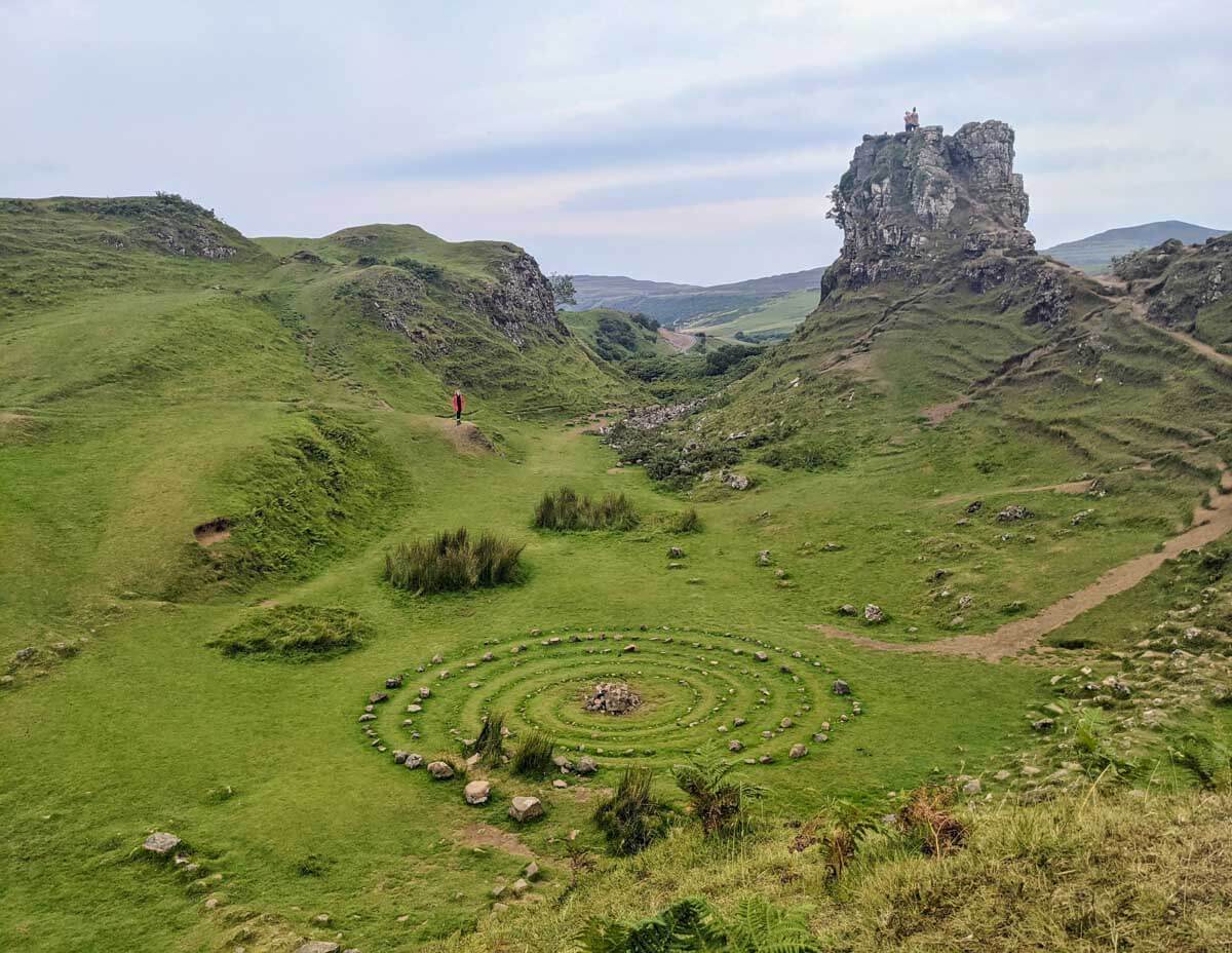 A lush, green valley known as Fairy Glen on the Isle of Skye, with rolling hills and a distinctive spiral of stones on the valley floor. A singular figure in red provides a stark contrast to the natural hues as they walk a path toward a rugged, towering rock formation with two people standing atop.