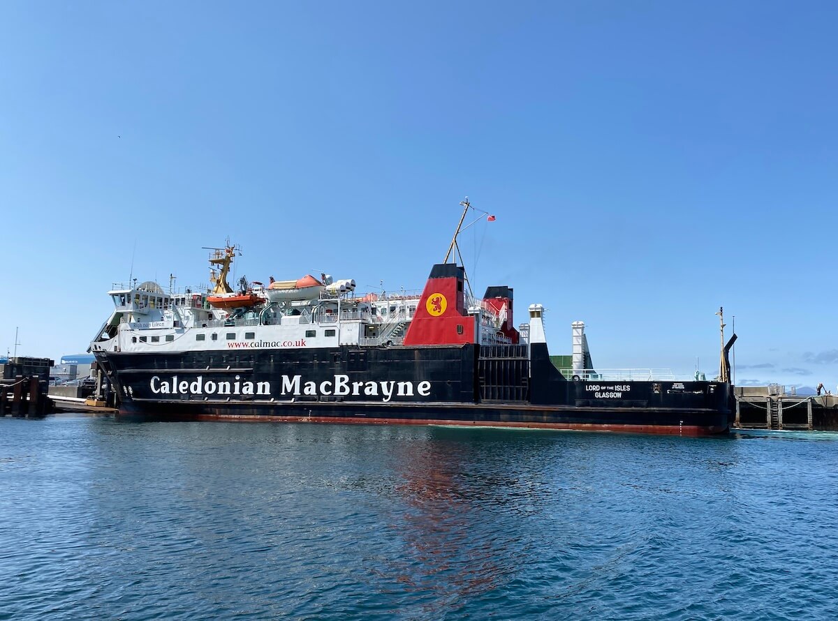 The 'Lord of the Isles' ferry, operated by Caledonian MacBrayne, is docked at Mallaig harbour on a bright sunny day. The vessel's striking black hull with the company logo is visible against the clear blue waters, while the red and black funnel proudly displays the company's lion rampant emblem