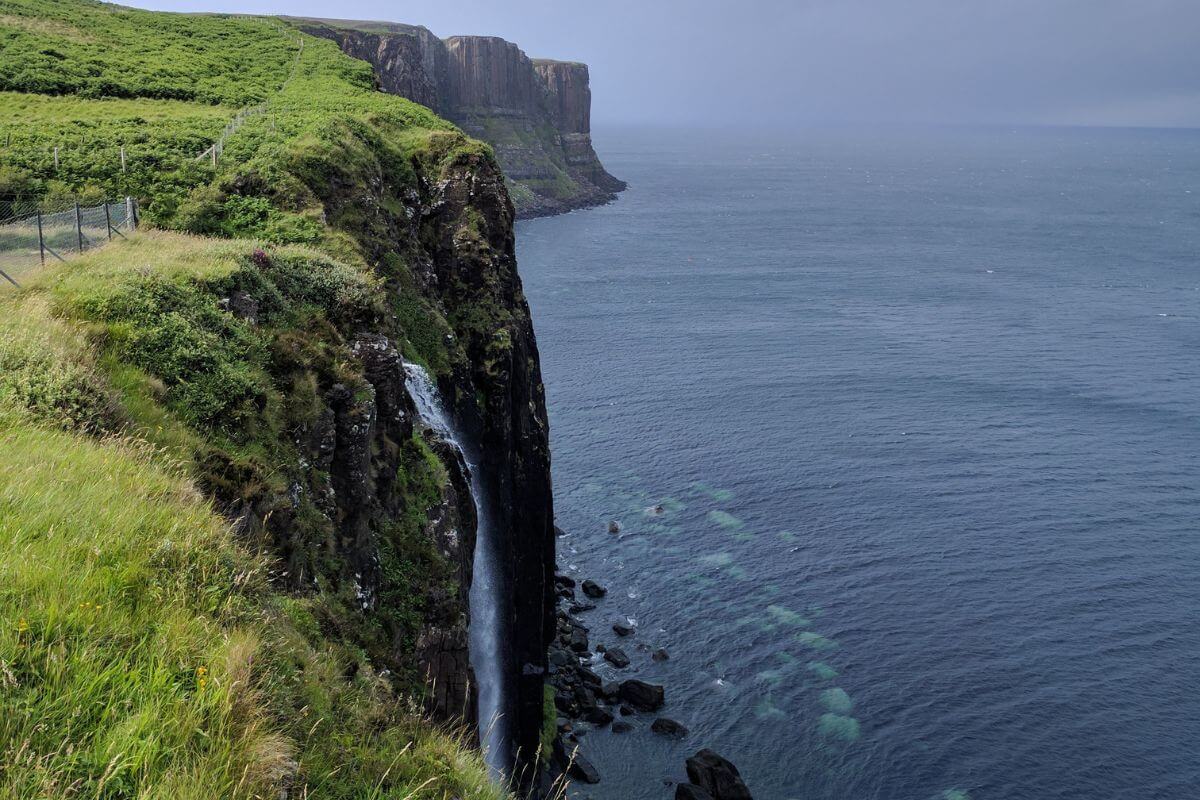 Mealt Falls on the Isle of Skye, where a slender waterfall cascades directly into the sea from a lush, green cliffside. The cliffs stretch into the distance, framing the vast ocean, under a grey, overcast sky, with faint patterns of underwater rocks visible through the clear water near the shore