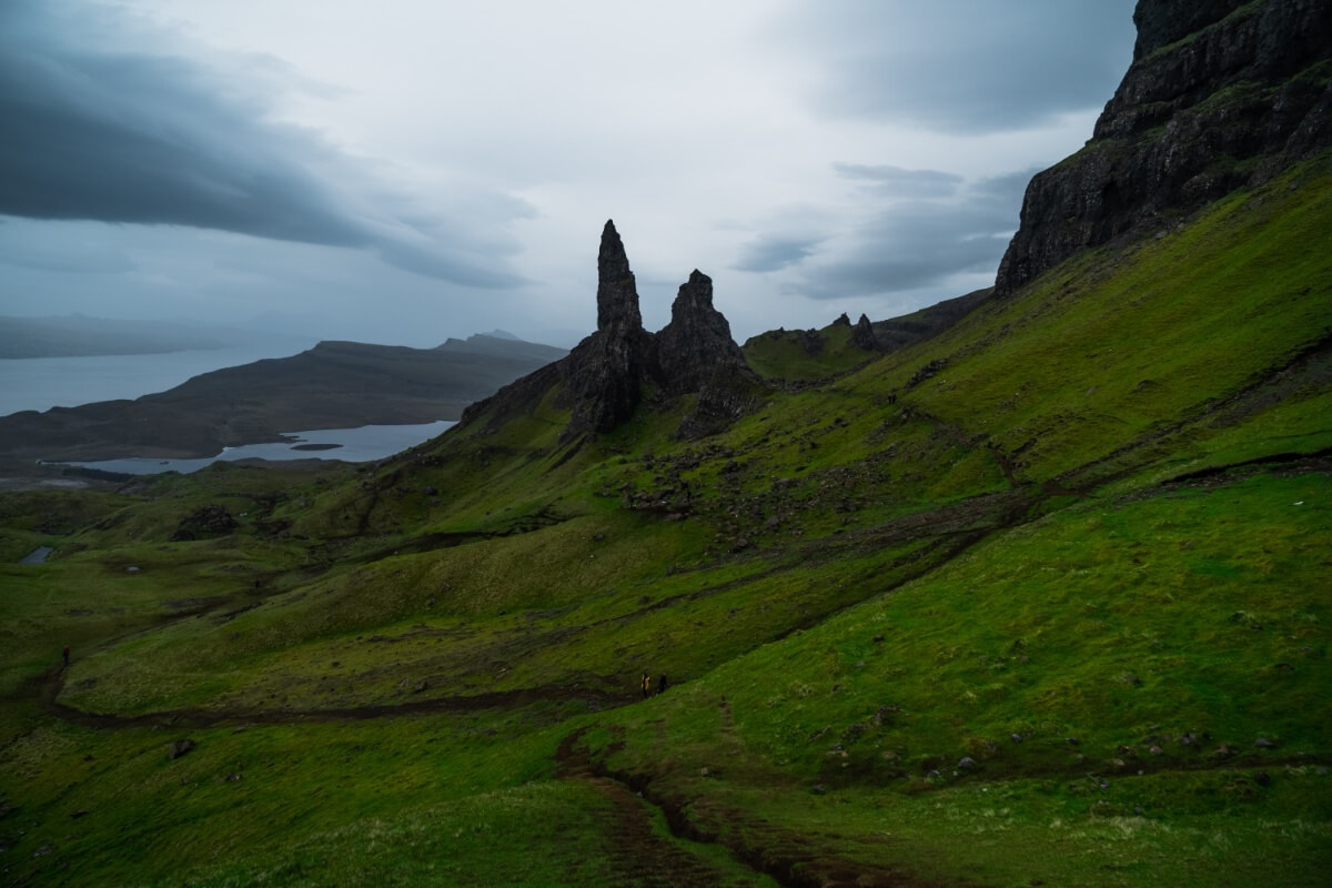 The iconic Old Man of Storr rock formations stand tall against a dramatic and moody sky on the Isle of Skye. Verdant slopes stretch around the imposing rocks, with a view of the sea in the distance, as winding trails lead towards the stark silhouettes of the ancient, jagged pinnacles
