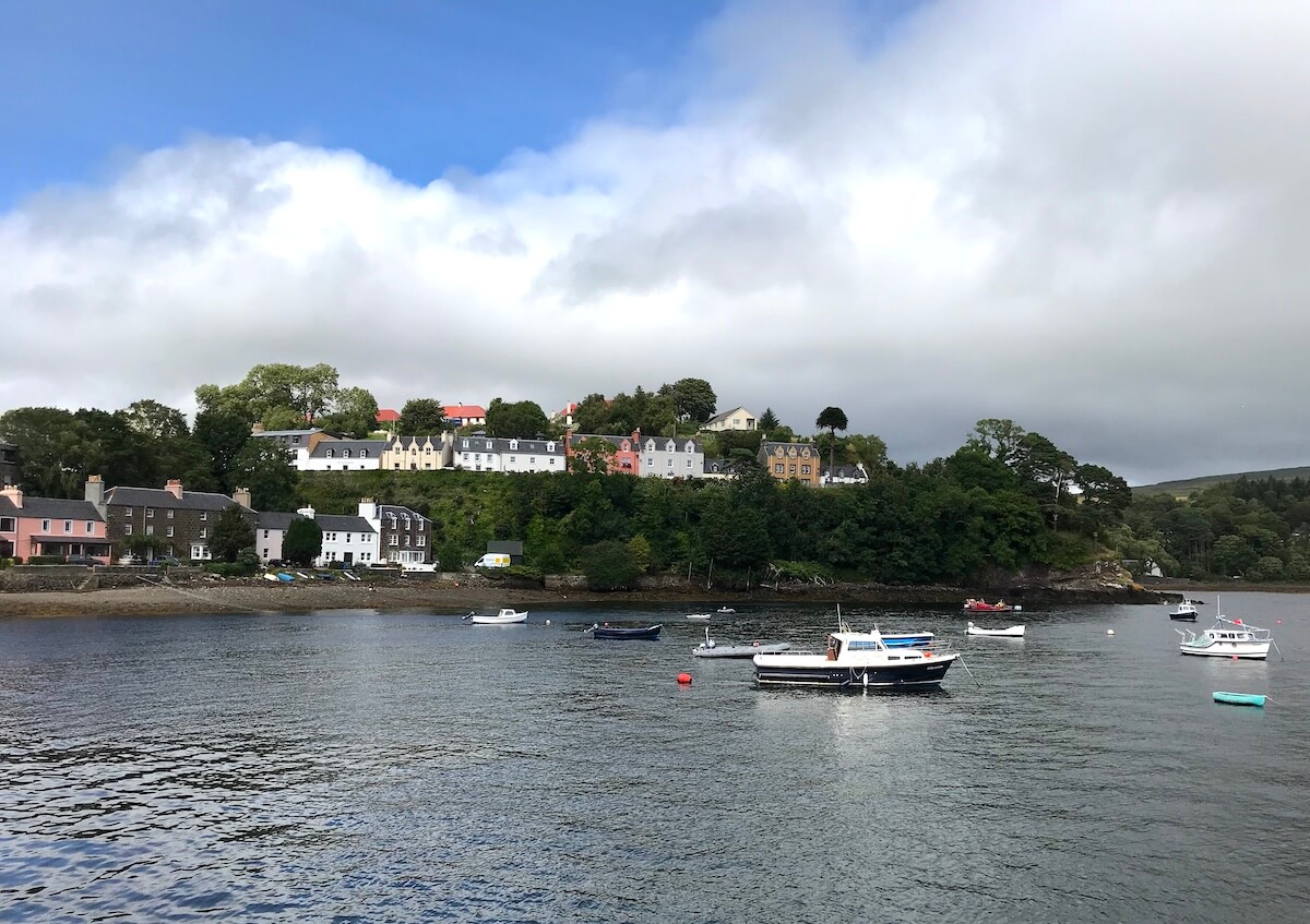 A serene harbour view of Portree, Isle of Skye, featuring a calm bay with boats anchored in the water. The shoreline is adorned with a quaint row of houses, painted in soft pastel colours, nestled among the trees and set against a backdrop of rolling hills and a cloudy sky