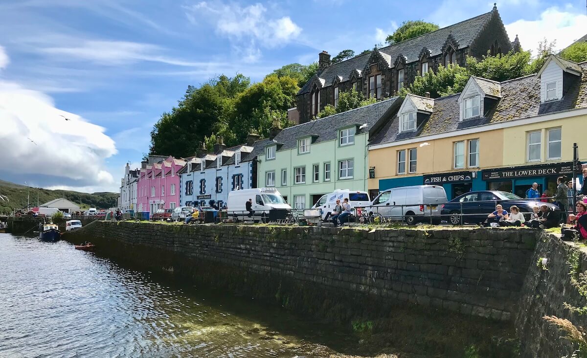A vibrant quayside scene in Portree, Isle of Skye, with a row of colourful houses in shades of pink, blue, and yellow. People are enjoying the waterfront, sitting on the sea wall, with a clear view of the calm bay waters, boats, and a bustling atmosphere against a backdrop of green hills and a bright blue sky.