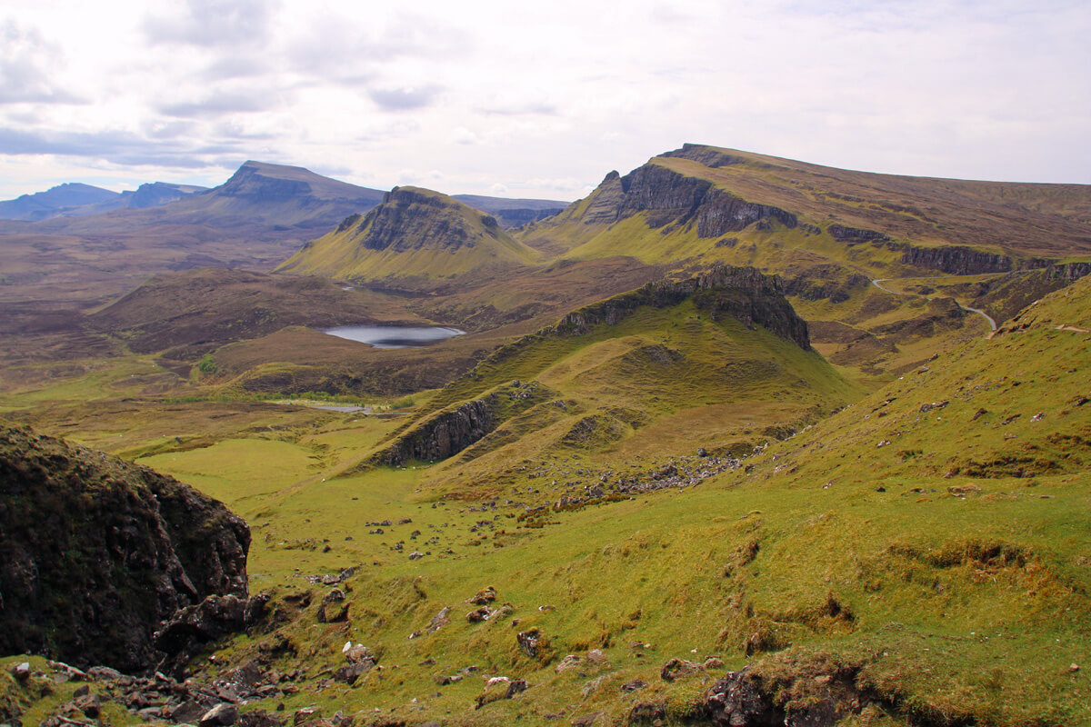 A sweeping view of the Quiraing landscape on the Isle of Skye, with its distinctive rolling hills, craggy cliffs, and a serene loch nestled in the valley. The green hues of the grassy terrain are set against a soft, cloudy sky, conveying the tranquil and rugged beauty of the Scottish Highlands