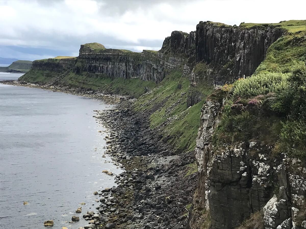 The rugged coast near Kilt Rock on the Isle of Skye, with towering basalt cliffs dropping steeply to a stony beach below. Lush green vegetation caps the clifftops, adding a dash of colour to the dramatic landscape, all under a subdued sky with the sea stretching into the distance