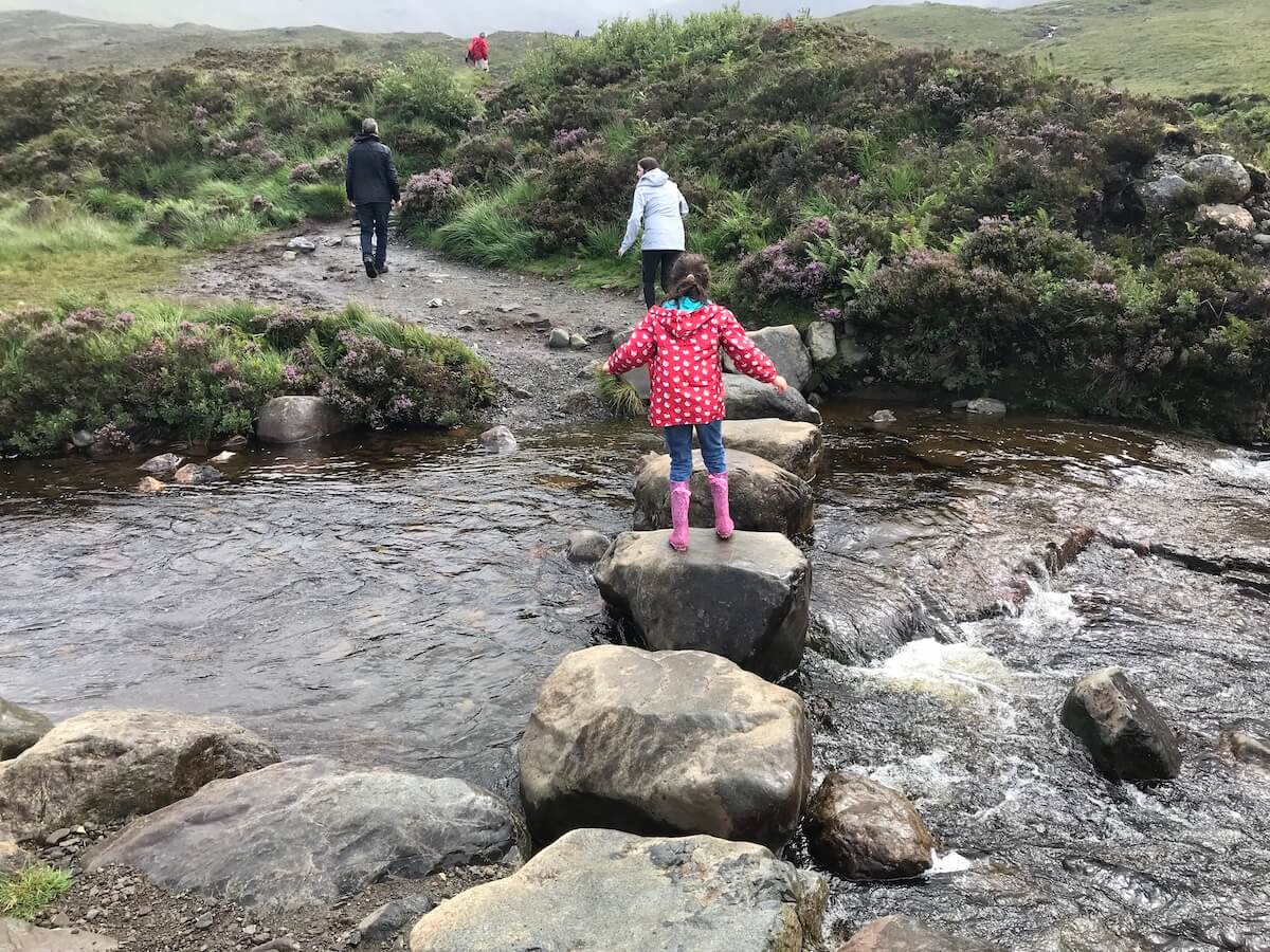 Author's young daughter in a red polka-dot coat and pink wellies balances on stepping stones to cross a stream on the trail to the Fairy Pools, Isle of Skye. Heather and greenery flank the brook, with the adults in the background walking along the path, all set in a serene moorland landscape