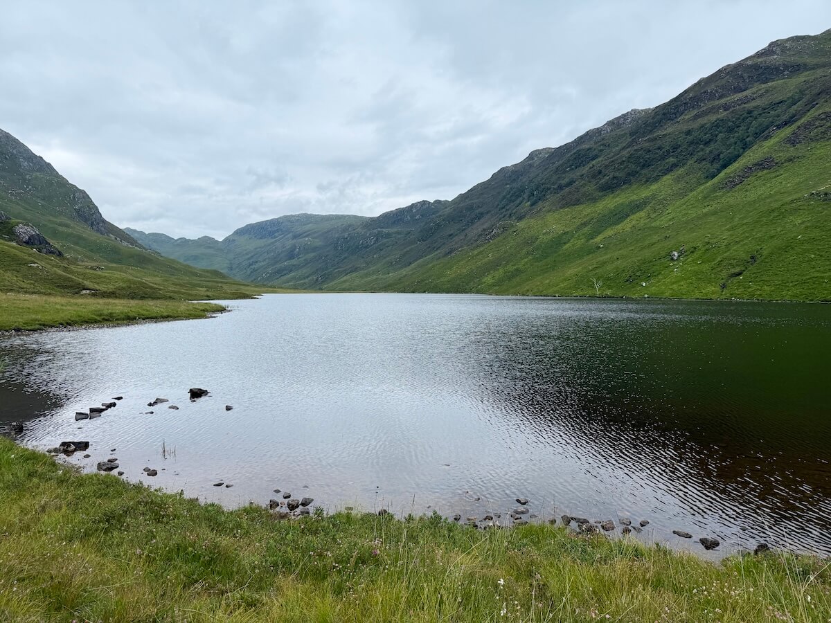 A calm freshwater loch nestled between rolling green hills under a cloudy sky. The still water reflects the moody light, with stones dotting the grassy shoreline.