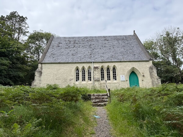 A small, cream-coloured stone church with a steep slate roof and arched windows, set among lush greenery with a narrow gravel path leading to its bright turquoise door.