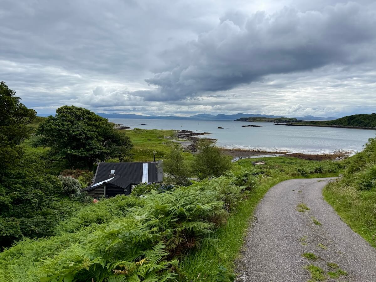Winding single-track road leading down to a rocky coastline with a black-roofed cottage nestled among trees and ferns, looking out over calm sea waters and distant hills.