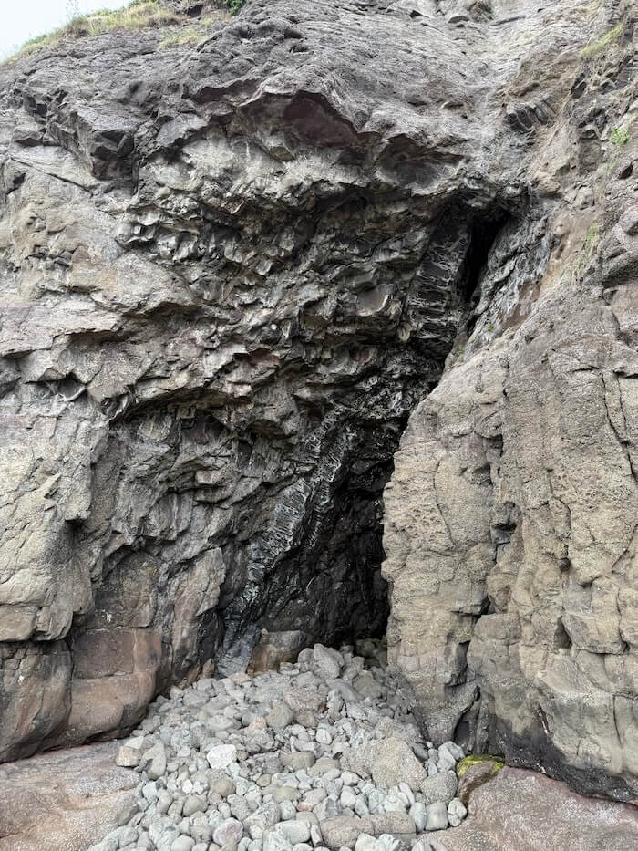 Steep rocky cliff face with a narrow cave entrance partially hidden among jagged stone and loose rocks at the base. The rough layers of rock frame the opening.
