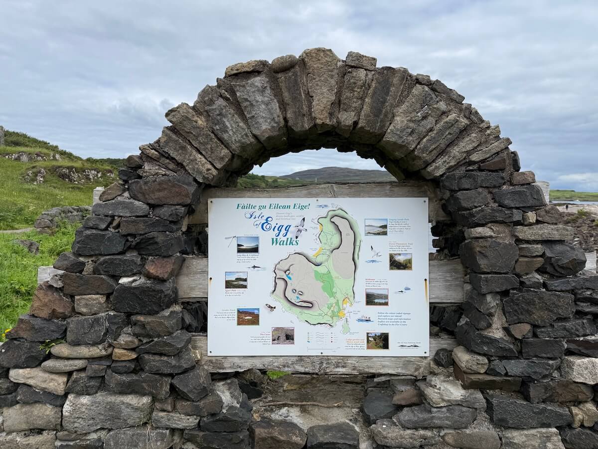 Stone arch framing an information board showing a map of walking routes on the Isle of Eigg, with grassy hills and a cloudy sky in the background.