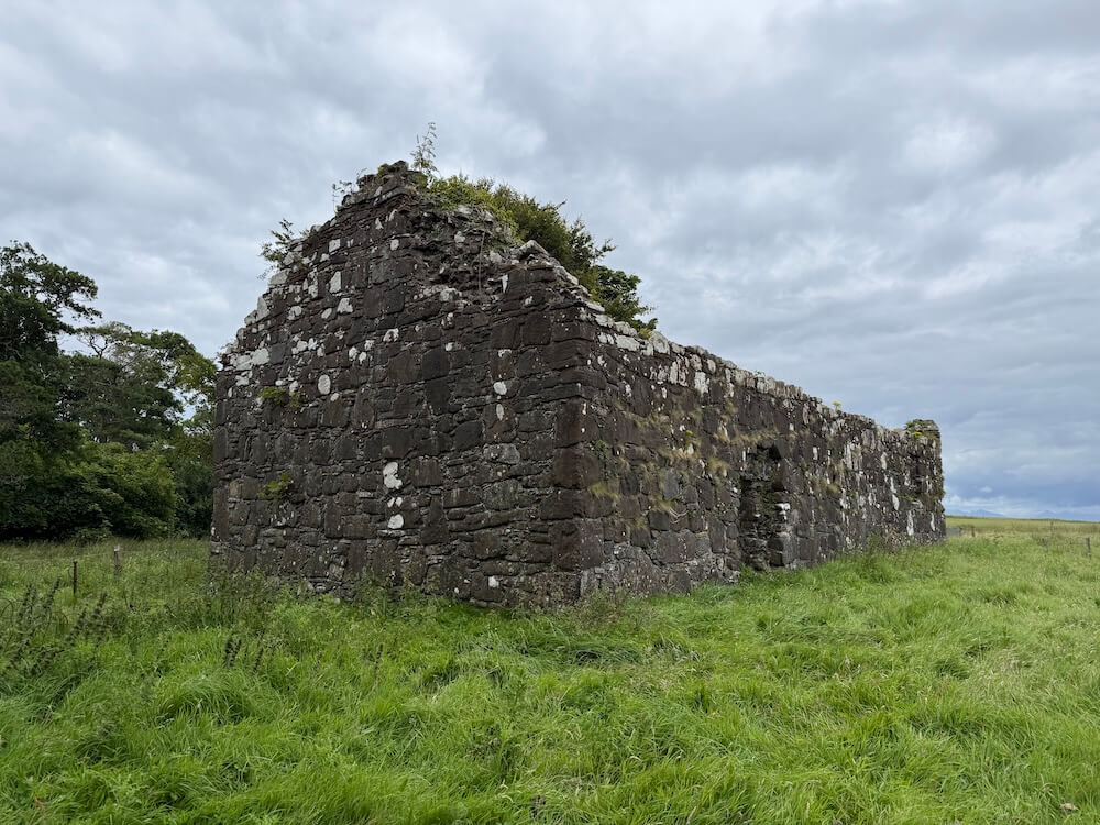 A weathered stone building stands alone in a grassy field, its roof missing and patches of white lichen scattered across the dark stone walls. The structure appears partially collapsed, surrounded by open countryside beneath a grey sky.