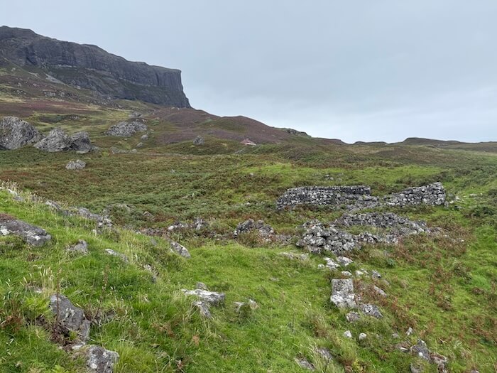 Low stone ruins sit scattered across a grassy hillside, with a steep rocky cliff rising in the background. The landscape appears windswept under a grey sky.