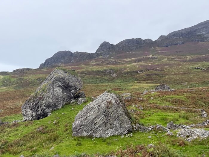 Large weathered boulders rest on green moorland, with rolling hills and rugged ridges stretching behind them. The scene feels open and exposed beneath cloud cover.