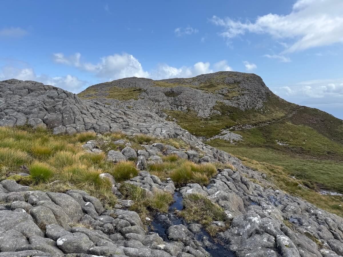Rocky hillside with patches of grass and small pools of water slopes up towards a rounded ridge under a bright blue sky. The terrain appears uneven and exposed.
