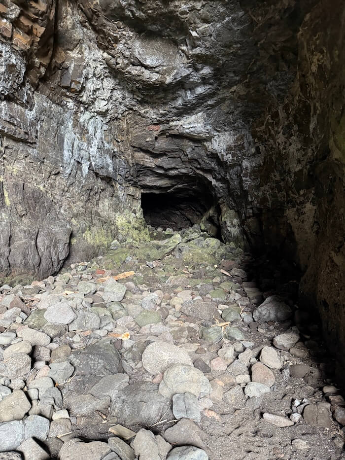 Wide cave interior with a rocky floor leads towards a dark tunnel opening in the far wall. The rough stone walls and scattered rocks create a rugged, enclosed space.