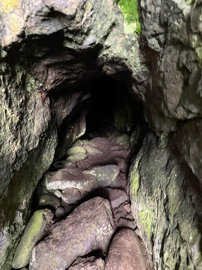 Narrow rocky opening at the back of a cave, with uneven stones on the ground leading into a darker tunnel. The tight passage suggests it opens into a larger space beyond.