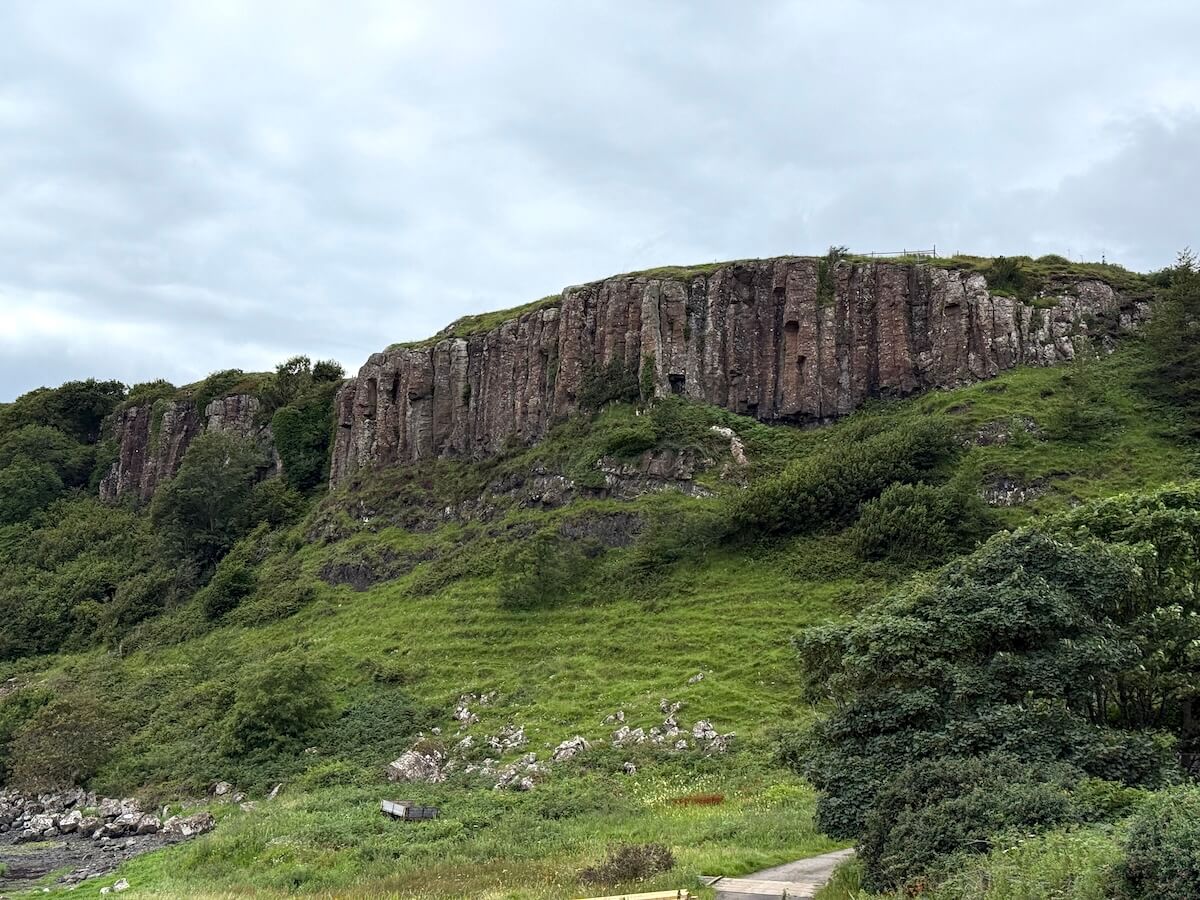 Steep grassy hillside rises to a ridge of tall, vertical rock formations, with patches of shrubs and trees scattered along the slope. A narrow path curves through the foreground beneath an overcast sky, highlighting the rugged landscape.