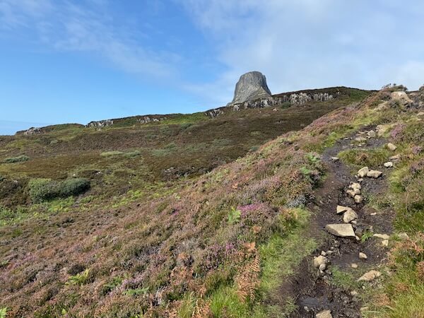 Narrow path climbs a hillside covered in heather and grass, leading towards a steep rocky peak in the distance. The trail looks rough and slightly muddy.