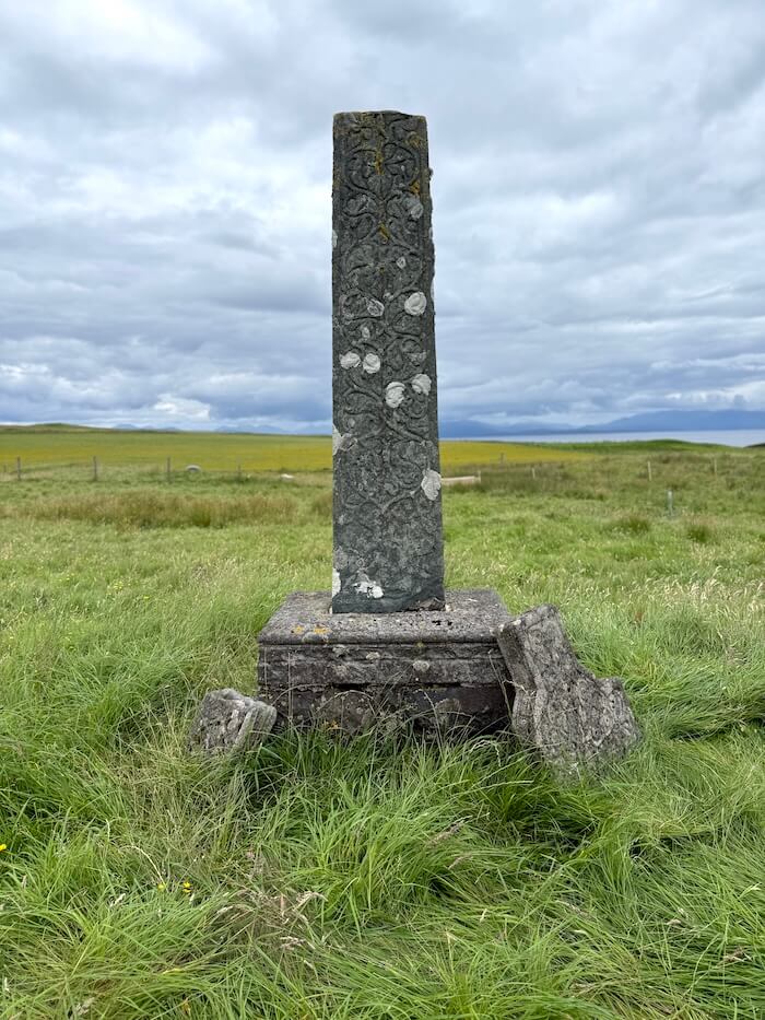 Tall carved stone pillar stands in a grassy field, decorated with worn patterns and patches of lichen, with a low horizon and distant hills under a cloudy sky. Smaller broken stones rest at its base, suggesting an old monument or grave marker.