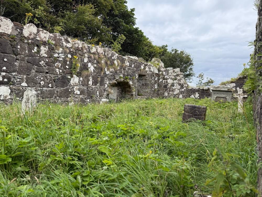 Overgrown grass fills the centre of a roofless stone ruin, with moss and lichen covering the old walls and a small gravestone visible among the greenery. The scene feels quiet and abandoned under a cloudy sky.