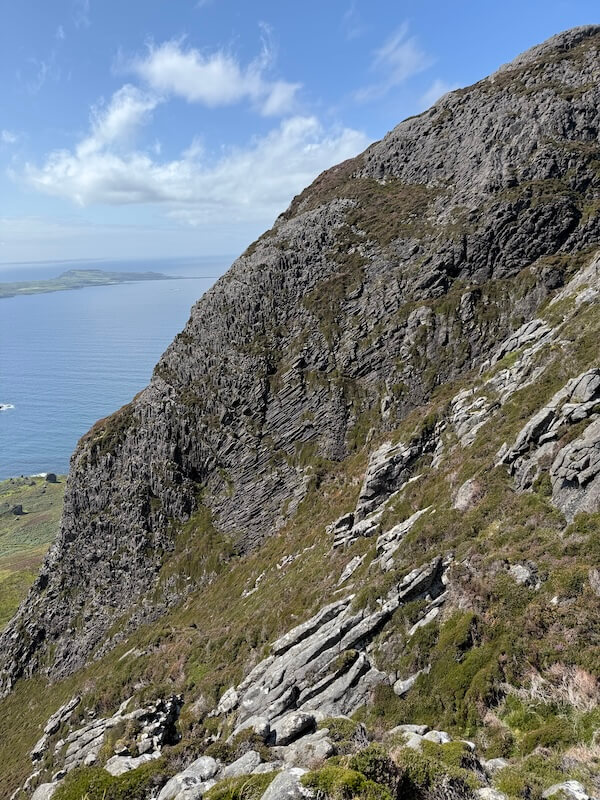 Steep rocky cliff rises above the sea, with layered stone and patches of grass clinging to the slope. The coastline and distant land are visible below under a bright sky.
