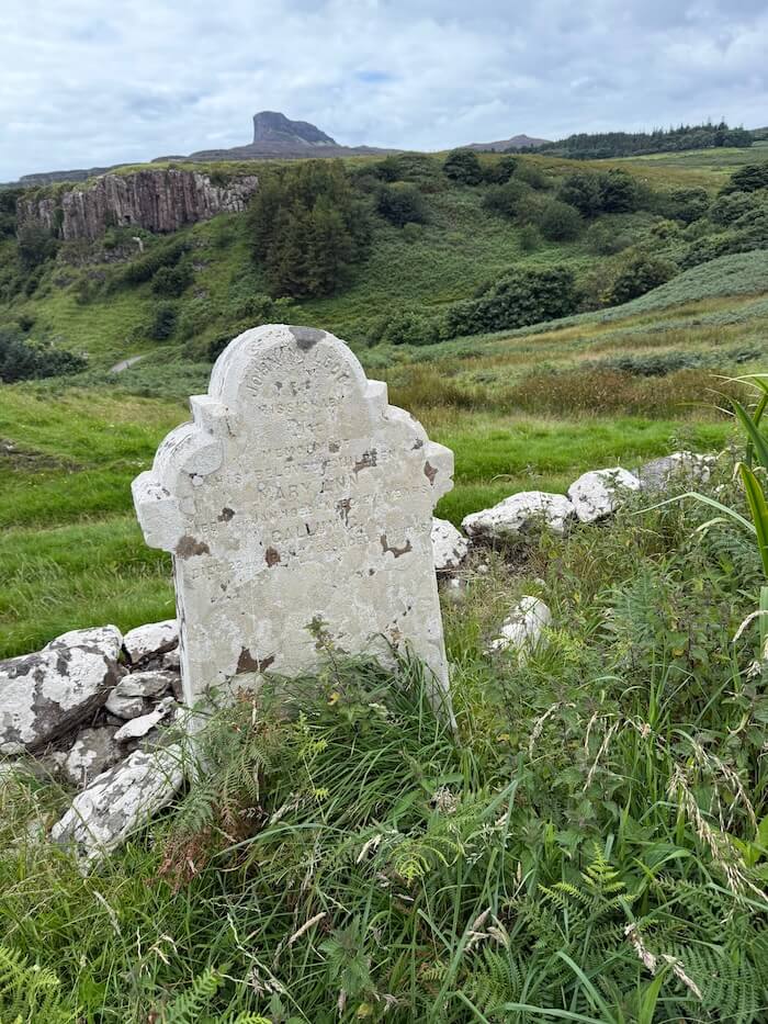 Weathered gravestone leans slightly among tall grass and ferns, with faint engraved text barely visible on its surface. In the background, green hills rise towards basalt column cliffs and a distant peak beneath a grey sky.