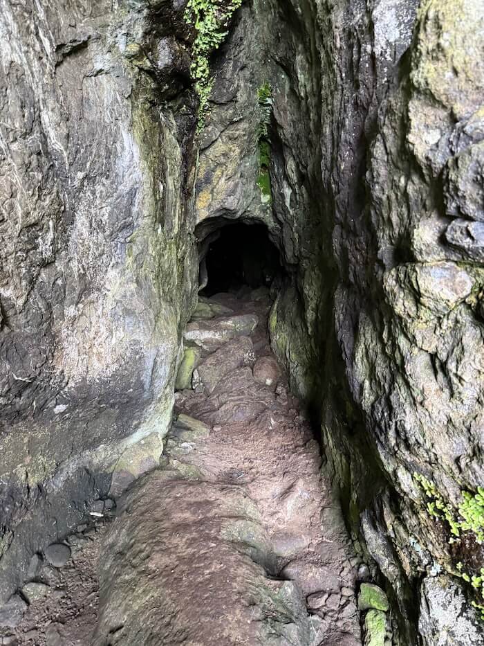 Tight rocky passage inside the cave shows rough walls and a narrow floor of stones leading into darkness. The space appears enclosed and slightly damp.