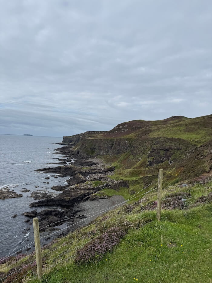 Rocky coastline curves along the water, with grassy cliffs above and waves breaking against dark stones. A wire fence runs along the edge of the slope.