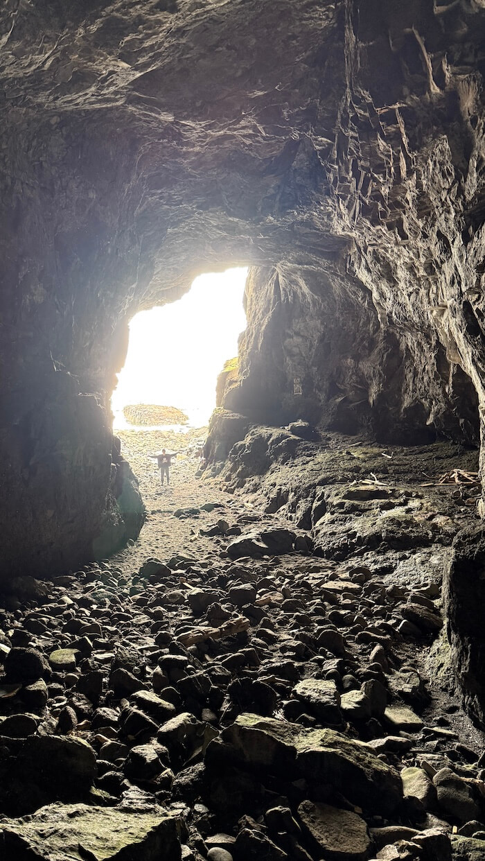 Large cave opens towards the sea, with light pouring in and a small figure standing near the entrance on a rocky floor. The bright opening contrasts with the dark interior.