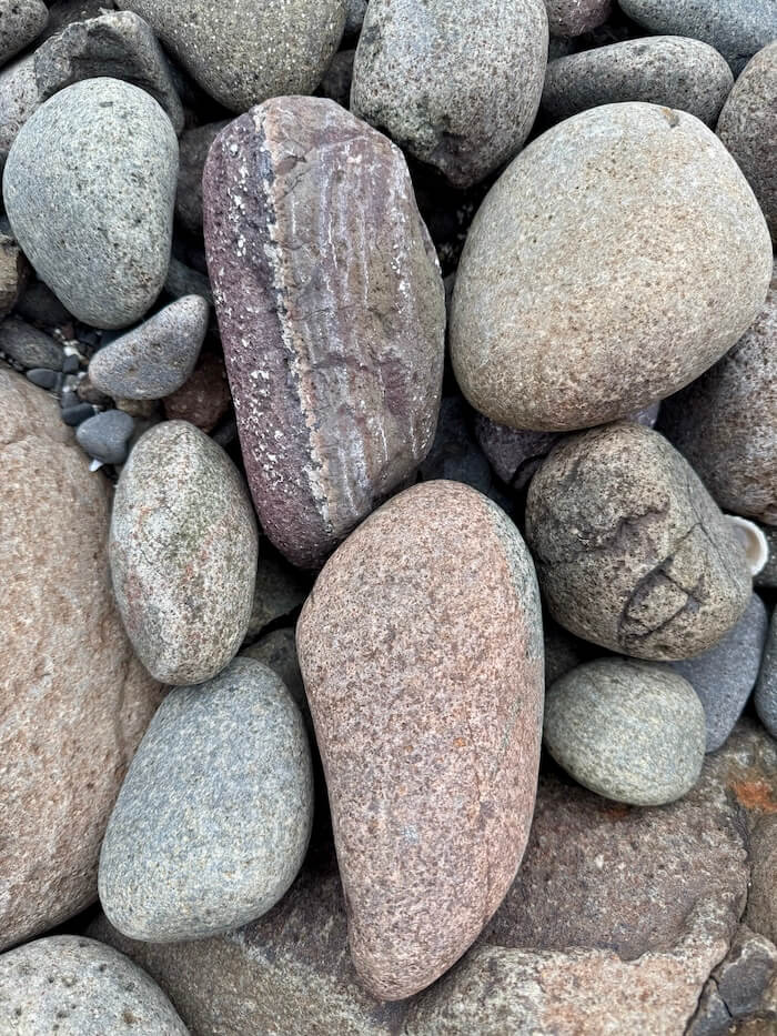 Collection of rounded beach stones in shades of grey and pink fills the frame. One elongated stone with layered stripes lies near the centre.