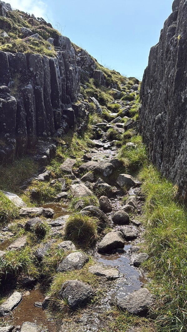 Steep stony path runs between tall rock walls, with small puddles and uneven footing along the way. The enclosed passage leads uphill.