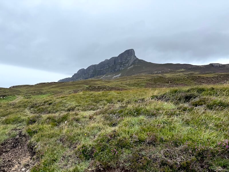 Grassy moorland stretches towards a steep, jagged peak under low cloud. A faint path cuts through the foreground across the open terrain.