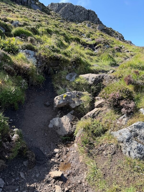 Rough dirt path cuts through grassy slopes dotted with rocks and heather, leading towards jagged cliffs above. The climb appears uneven and steep.