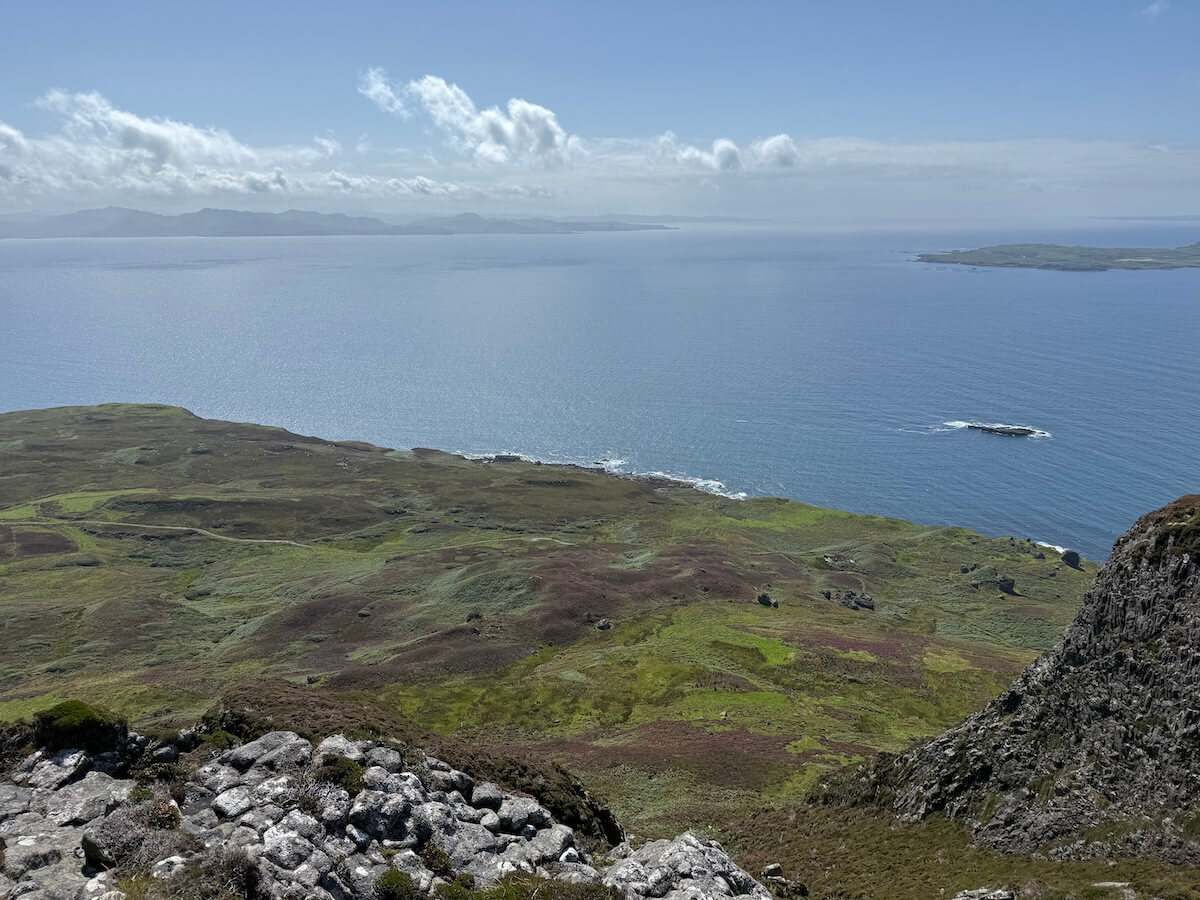 High viewpoint looks out over green coastal land and calm sea, with distant islands visible on the horizon. Sunlight reflects off the water beneath scattered clouds.